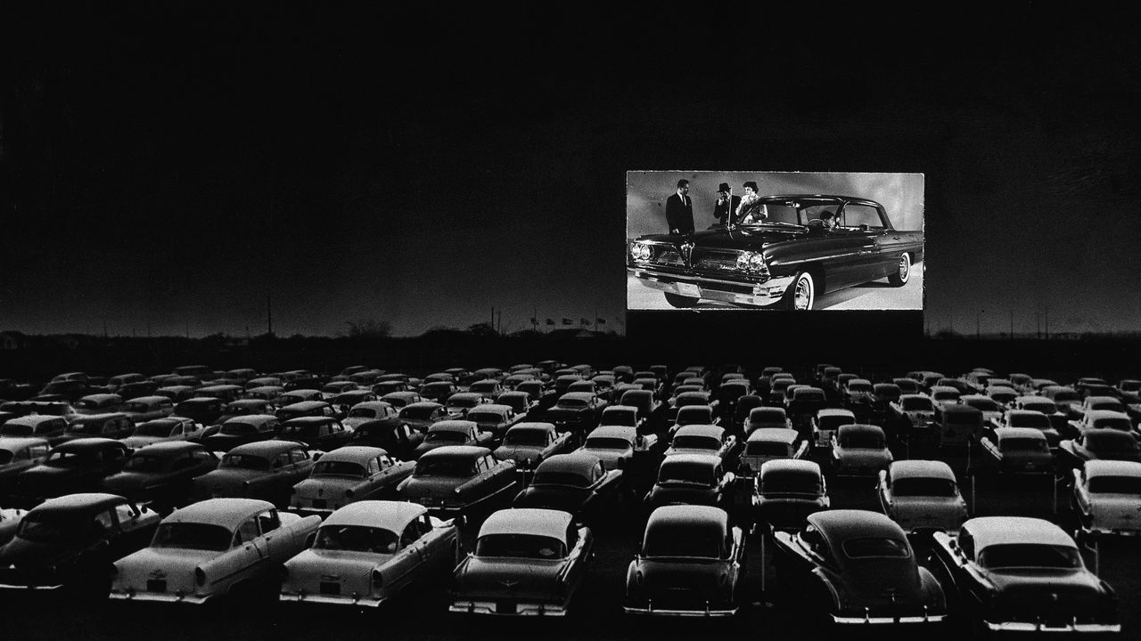 Vehicles fill a drive-in theater while people on the screen stand near a new car, 1950s.