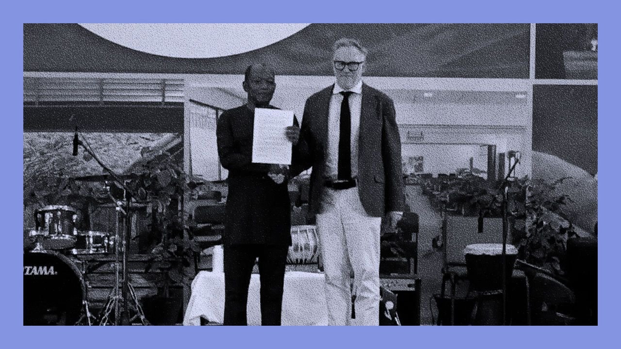 Charlie Gladstone and Eric Philips stand on the stage at the University of Guyana holding up an apology letter signed by the Gladstone family for the actions of their ancestor, Sir John Gladstone